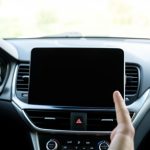 A car dashboard with a dark, blank center infotainment screen and a hand reaching toward it